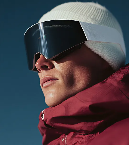 Person wearing a white beanie, reflective ski goggles, and a maroon jacket against a dark blue sky.