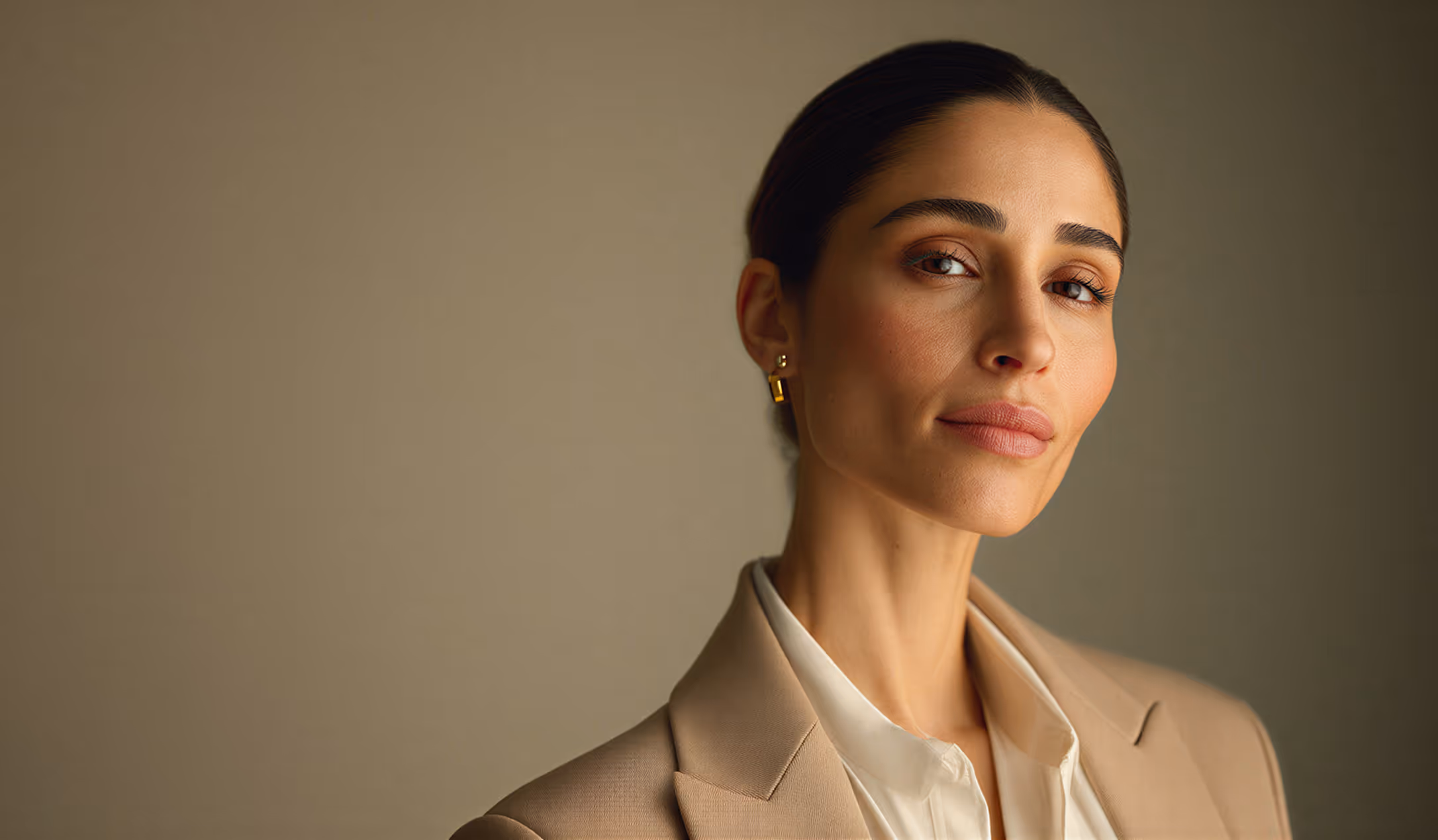 Confident woman with slicked-back hair wearing a beige blazer and white blouse against a plain background.