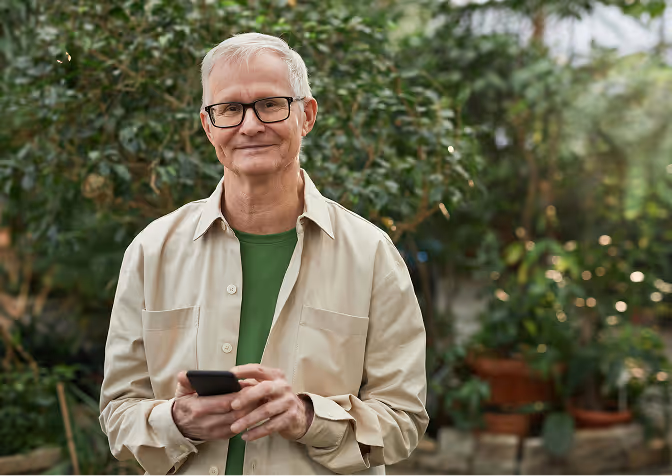 Smiling elderly man with glasses holding a smartphone in a garden with green plants in the background.