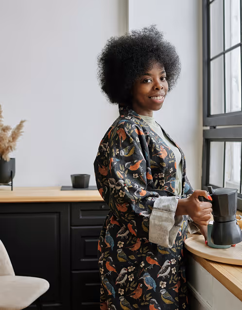 Smiling woman in a bird-patterned robe preparing coffee by a window in a modern kitchen.
