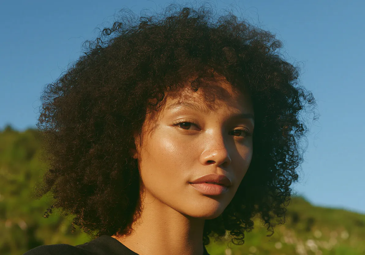 Close-up of a young woman with curly hair, natural makeup, and a serene expression against a blue sky and greenery background.