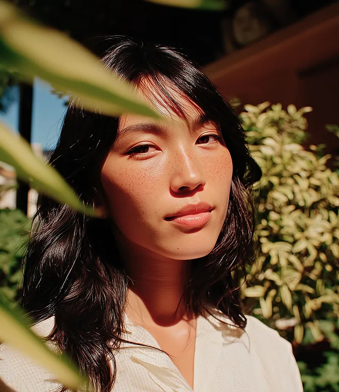 Close-up of a young woman with dark hair and freckles, standing outdoors with green foliage in the background.