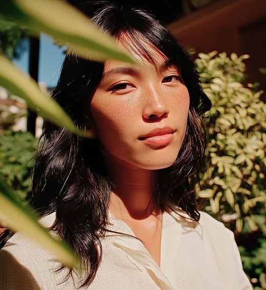 Portrait of a woman with long dark hair and freckles, standing outdoors with green plants in the background.