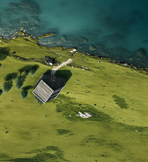 Aerial view of a small wooden cabin casting a shadow on green grassy land beside a rocky shoreline with clear blue water.