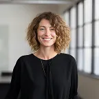 Smiling woman with curly hair wearing a black top in a bright office space with large windows.