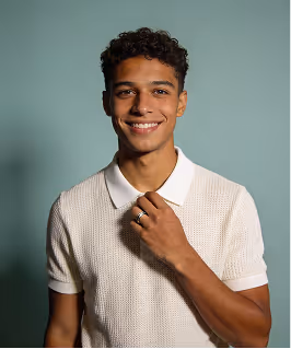 Young man with curly hair smiling, wearing a white collared knit shirt against a gray background.