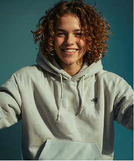 Smiling person with curly hair wearing a light gray hoodie against a dark green background.