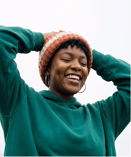Smiling woman in a green hoodie and orange knit hat with hands behind her head.