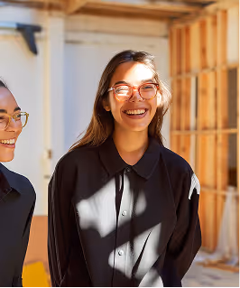 Two smiling women wearing glasses in a sunlit indoor setting with wooden beams in the background.