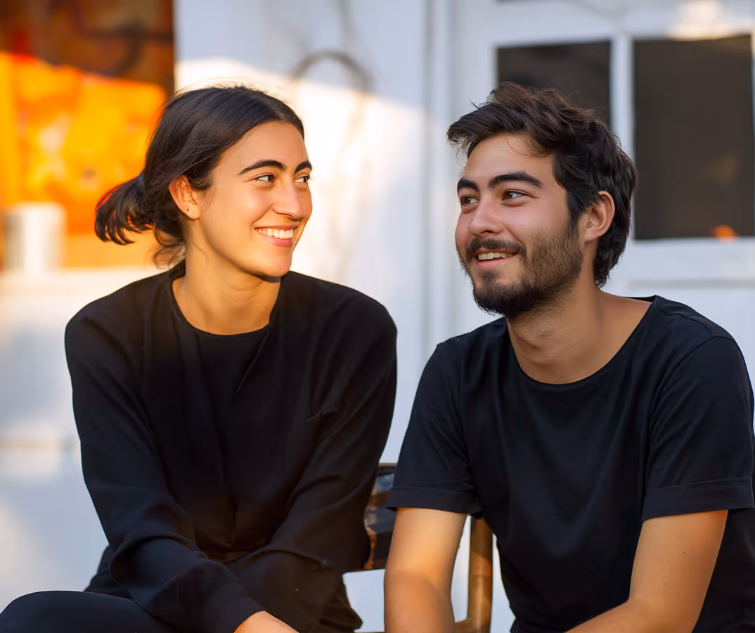Smiling young woman and man sitting next to each other, both wearing black shirts, in a well-lit outdoor setting.