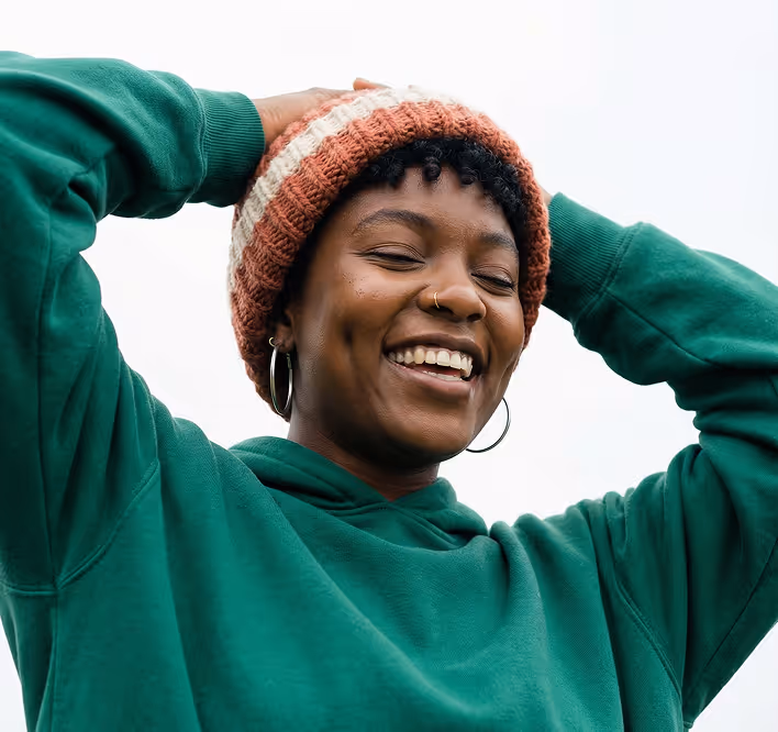 Smiling person wearing a green hoodie and a knitted hat with hands touching the hat.